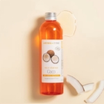 Un flacon transparent d'Aroma Zone Huile de Coco BIO 100ml repose sur une surface beige avec un petit déversement au sommet, à côté de deux morceaux de noix de coco et de son étiquette ornée d'images de noix de coco. Dakar