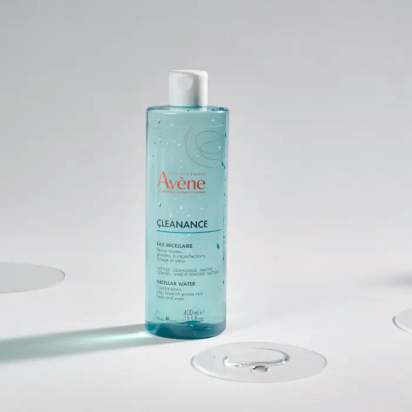 Un flacon bleu d'Eau Micellaire Avène Cleanance est posé debout sur une surface claire. Transparent, il laisse apparaître l'eau qu'il contient et possède un bouchon blanc. Quelques gouttes et des marques circulaires à proximité suggèrent l'utilisation du produit. Dakar