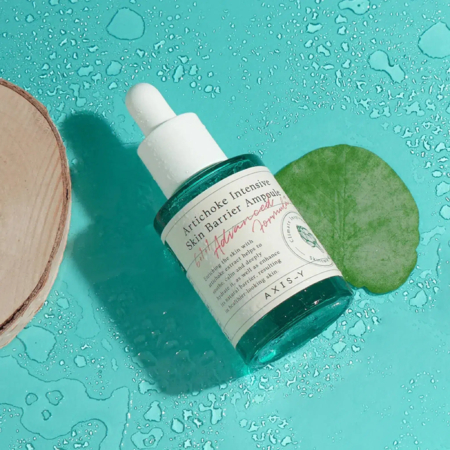 Un flacon d'ampoule Artichaut Barrière Intensive pour la Peau, muni d'un bouchon compte-gouttes, repose sur une surface bleu-vert humide, à côté d'une feuille verte et d'un morceau de bois. Des gouttes d'eau s'éparpillent. Dakar