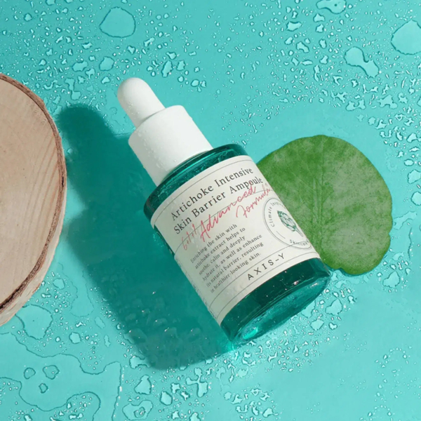 Un flacon d'ampoule Artichaut Barrière Intensive pour la Peau, muni d'un bouchon compte-gouttes, repose sur une surface bleu-vert humide, à côté d'une feuille verte et d'un morceau de bois. Des gouttes d'eau s'éparpillent. Dakar