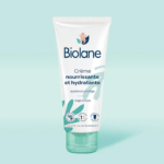 Un tube de crème hydratante Biolane est présenté sur un fond bleu sarcelle clair. L'étiquette met en avant ses propriétés nourrissantes et hydratantes, avec des pictogrammes indiquant 98 % d'origine naturelle et des tests dermatologiques. Dakar