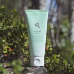 Un tube de nettoyant rafraîchissant à la prune verte Beauty of Joseon est posé sur un rocher en plein air, entouré d'une végétation floue. L'emballage vert présente des textes en anglais et en coréen. La lumière du soleil met le produit en valeur, soulignant le cadre naturel. Dakar