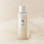 Une bouteille en verre dépoli avec un bouchon blanc, étiquetée « Glow-Repairing Face Milk » en caractères anglais et coréens, placée sur un fond en tissu doux de couleur crème. Dakar