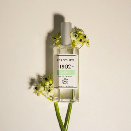 Un flacon d'Eau de Cologne Jasmin Berdoues 1902 se dresse droit, entouré de délicates fleurs blanches. Le flacon est transparent, avec une étiquette blanche et verte et un bouchon blanc. Le fond clair rehausse l'élégance de la composition. Dakar
