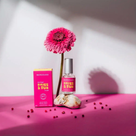 Un flacon de parfum rose étiqueté « Baies & Fun » repose à côté de sa boîte sur une surface magenta. Un gerbera rose se dresse derrière, émergeant d'un galet. De petites perles rouges sont dispersées au premier plan. Dakar
