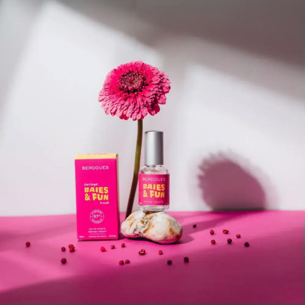 Un flacon de parfum rose étiqueté « Baies & Fun » repose à côté de sa boîte sur une surface magenta. Un gerbera rose se dresse derrière, émergeant d'un galet. De petites perles rouges sont dispersées au premier plan. Dakar