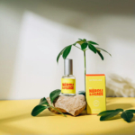 Un vaporisateur et une boîte étiquetés « Néroli et Paix » sont posés sur une surface jaune. Une petite plante en pot et une pierre lisse avec des feuilles sont disposées à proximité, ajoutant une touche naturelle à la composition. Les ombres créent une ambiance apaisante. Dakar