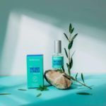 Un flacon de parfum Berdoues « Le Vert & Libre » est posé sur une surface turquoise, à côté de son écrin. Une petite branche aux feuilles vertes et un rocher se trouvent à côté du flacon, créant une ambiance naturelle et fraîche. Dakar