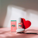 Un petit flacon de parfum étiqueté « Osmanthus & Love » est posé sur une pierre lisse, à côté d'une boîte rose assortie. Un cœur rouge s'appuie contre le flacon, baigné d'une douce lumière naturelle. Les ombres créent une ambiance chaleureuse et accueillante. Dakar