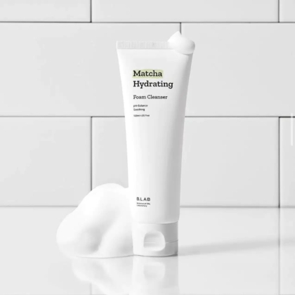Un tube de mousse nettoyante hydratante Matcha est posé sur une surface carrelée blanche. Blanc, il est orné d'un texte noir et vert, et la mousse est disposée avec art à sa base et à son sommet, créant un look épuré et minimaliste. Dakar