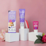 Un kit d'épilation comprenant trois produits : un coffret, un tube de crème dépilatoire et une crème de soin apaisante. Ils sont présentés sur des supports blancs ornés de roses roses et d'une éponge sur fond rose. Dakar