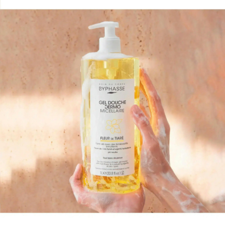 Des mains couvertes de mousse savonneuse tiennent un grand flacon transparent de Gel Douche Dermo Micellaire Byphasse contenant un liquide jaune. L'étiquette porte la mention « Fleur de Tiaré ». Le fond est une surface texturée marron clair. Dakar