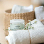 Un pot de masque exfoliant Candès Éclat est posé sur une serviette blanche roulée, ornée de petites fleurs blanches. En arrière-plan, un panier tressé accueille des serviettes similaires, créant une ambiance de spa. Dakar