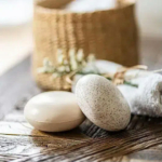 Deux savons ovales, l'un lisse, l'autre moucheté, reposent sur une surface tissée. Une serviette blanche roulée et un panier en osier beige orné de fleurs blanches se détachent en arrière-plan flou, créant une ambiance de spa. Dakar