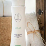 Un flacon de tonique éclaircissant Candes Éclat est posé à côté d'une serviette blanche enroulée et nouée avec de la ficelle. Le flacon est blanc avec des détails argentés, et la serviette est posée sur un plateau en bois naturel et rustique. Dakar