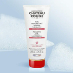 Un tube de Gel Nettoyant Moussant Laboratoire Château Rouge est centré sur un fond bleu clair orné de bulles de mousse. Blanc, il est rehaussé de touches de rouge et porte l'inscription « Teint Éclat ». Son poids net est de 200 ml. Dakar