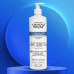 Un flacon de Lait de Beauté Ultra Hydratant Château Rouge est posé sur une plateforme circulaire bleue. Le flacon pompe blanc est étiqueté en français et en anglais, soulignant ses propriétés pour les peaux très sèches et une hydratation 24 heures sur 24. Le fond est bleu et abstrait. Dakar