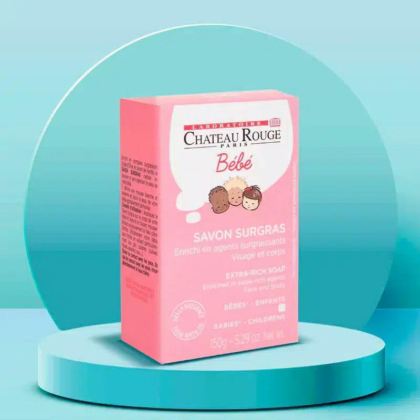 L'image montre une boîte rose de savon extra-riche Château Rouge Paris Bébé pour bébés et enfants, présentée sur une plateforme circulaire bleu sarcelle. La boîte présente des visages d'enfants dessinés et un texte détaillant les informations produit. Le fond est un dégradé bleu sarcelle. Dakar