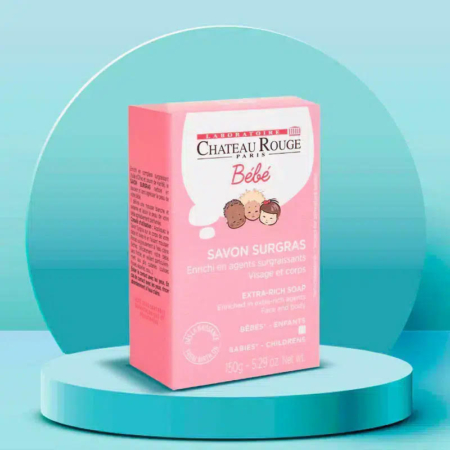 L'image montre une boîte rose de savon extra-riche Château Rouge Paris Bébé pour bébés et enfants, présentée sur une plateforme circulaire bleu sarcelle. La boîte présente des visages d'enfants dessinés et un texte détaillant les informations produit. Le fond est un dégradé bleu sarcelle. Dakar