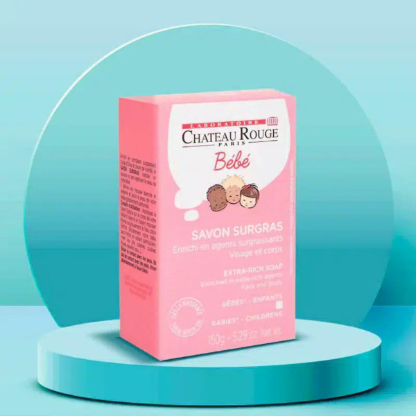 L'image montre une boîte rose de savon extra-riche Château Rouge Paris Bébé pour bébés et enfants, présentée sur une plateforme circulaire bleu sarcelle. La boîte présente des visages d'enfants dessinés et un texte détaillant les informations produit. Le fond est un dégradé bleu sarcelle. Dakar