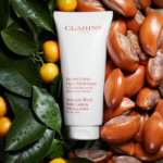 Un tube de Lait Corps Hydratant Clarins est entouré de noix de karité, de leurs coques craquelées et de feuilles vertes. Ce lait est indiqué pour les peaux très sèches et contient du beurre de karité. Dakar