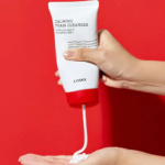 Une main distribue une mousse nettoyante blanche dans un tube étiqueté « Mousse Nettoyante Calmante » avec des touches de rouge sur fond rouge. Le tube est tenu au-dessus d'une paume ouverte et le produit est appliqué. Dakar