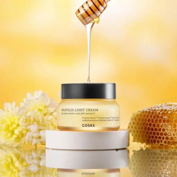Un pot de crème légère à la propolis COSRX posé sur un socle blanc. Du miel coule d'une louche à miel au-dessus, avec un rayon de miel et des fleurs blanches à côté. Le fond est un doux dégradé de jaune. Dakar