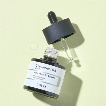 Un petit flacon foncé étiqueté « The Retinol 0.5 » de COSRX, avec un compte-gouttes noir posé sur le côté, sur fond vert clair. Quelques gouttes de produit sont visibles près du flacon. Dakar