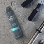 Une bombe de déodorant Dove Men+Care Clean Comfort repose sur une surface grise texturée. À proximité se trouvent les poignées d'une corde à sauter et d'une barre de musculation en métal, suggérant un style de vie actif ou de remise en forme. Dakar