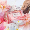Un flacon d'Eau de Parfum « Miss Dior » orné d'un nœud argenté trône au milieu de fleurs roses et blanches. Un morceau de bois flotté en arrière-plan crée une composition élégante et raffinée. Dakar
