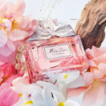 Un flacon d'Eau de Parfum « Miss Dior » orné d'un nœud argenté trône au milieu de fleurs roses et blanches. Un morceau de bois flotté en arrière-plan crée une composition élégante et raffinée. Dakar