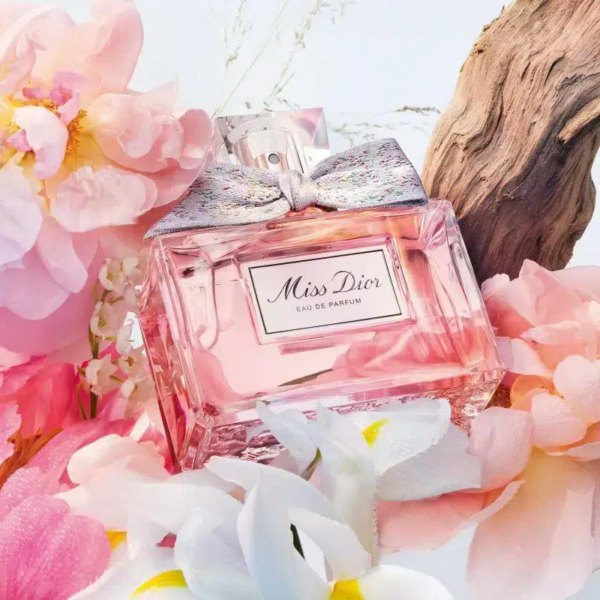 Un flacon d'Eau de Parfum « Miss Dior » orné d'un nœud argenté trône au milieu de fleurs roses et blanches. Un morceau de bois flotté en arrière-plan crée une composition élégante et raffinée. Dakar
