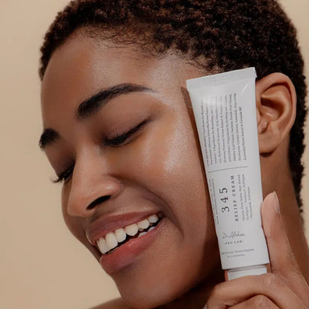 Une personne souriante, les yeux fermés, tient un tube de crème 345 Relief près de son visage. Aux cheveux courts et bouclés, elle applique la crème, révélant une peau lisse et éclatante. Dakar