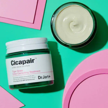 Un pot de traitement correcteur de couleur Dr. Jart+ Cicapair Tiger Grass, couvercle ouvert, repose sur une surface verte éclatante. La crème à l'intérieur est beige. Des formes géométriques roses sont dispersées, rehaussant l'esthétique lumineuse. Dakar