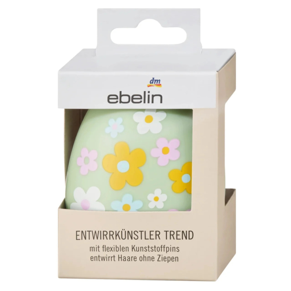 Une brosse à cheveux vert pastel présentée dans une boîte en carton. Elle présente un motif floral composé de fleurs jaunes, blanches, roses et violettes. L'emballage porte le logo de la marque « ebelin » et des informations produit en allemand. Dakar