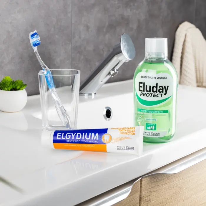 Une brosse à dents dans un verre transparent, un tube de dentifrice Elgydium et un flacon de bain de bouche Eluday trônent élégamment sur le lavabo blanc de la salle de bain. Un mur carrelé gris et une serviette contribuent à la sérénité du décor. Découvrez l'excellence des soins dentaires avec Univers Cosmetix. Dakar