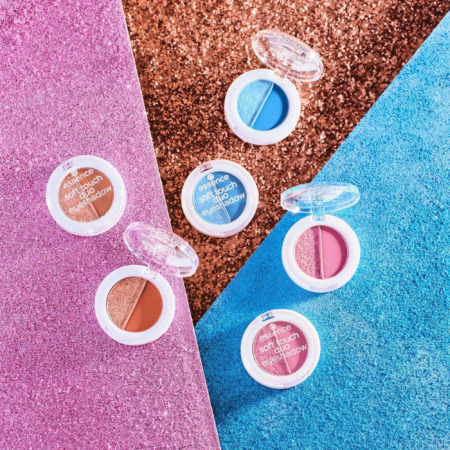 Six boîtiers de fards à paupières Essence Soft Touch sont disposés sur une surface texturée pailletée rose, bleue et cuivrée. Leurs teintes varient, notamment le bleu, le marron et le rose. Chaque boîtier est ouvert, laissant apparaître les couleurs des fards à paupières. Dakar