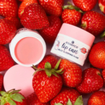 Un masque de nuit en gelée pour les lèvres, présenté dans un contenant rose, est entouré de fraises fraîches. Le pot ouvert, révélant le produit rose qu'il contient, se trouve à côté du pot fermé. La présentation met en valeur le thème de la fraise. Dakar