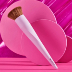 Un pinceau de maquillage avec un manche rose et des poils marron est présenté sur un fond vibrant de cercles roses superposés et d'une surface magenta brillante. Dakar
