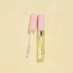 Un brillant à lèvres transparent avec un capuchon rose est ouvert, révélant la baguette applicatrice, sur un fond beige clair. Dakar