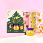 Coffret cadeau « Bonjour l'hiver » comprenant un baume à lèvres au miel Frudia et des crèmes pour les mains aux motifs fruités. Il comprend un petit pot et des tubes décorés de baies et de noix de coco. Des fleurs roses apparaissent en arrière-plan à droite. Dakar