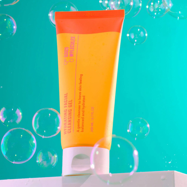 Un tube orange et jaune vif de Gel Nettoyant Hydratant Visage Good Molecules est posé sur une surface blanche. Des bulles de différentes tailles flottent autour, sur fond vert. Le texte sur le tube est partiellement visible. Dakar