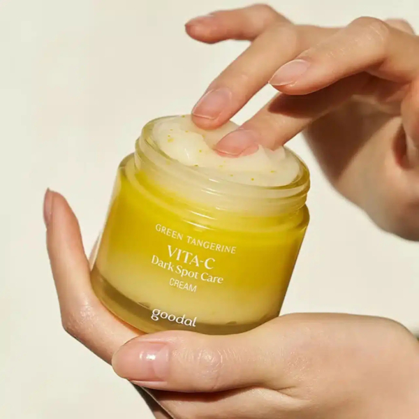 Des mains tiennent un pot ouvert de crème anti-taches Goodal Green Tangerine Vita-C. Une main trempe un doigt dans le produit crémeux, sous une lumière naturelle intense. Dakar