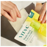 Des mains ouvrent un emballage étiqueté « Sérum anti-taches VITA-C » pour révéler un masque en tissu blanc. L'emballage est blanc et jaune avec un texte vert. Dakar