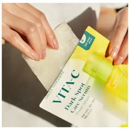 Des mains ouvrent un emballage étiqueté « Sérum anti-taches VITA-C » pour révéler un masque en tissu blanc. L'emballage est blanc et jaune avec un texte vert. Dakar