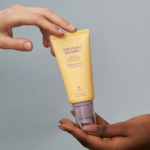 Deux mains tiennent un tube jaune de crème solaire Haruharu Wonder sur fond gris clair. Le tube porte un texte précisant sa protection SPF 50+. Dakar