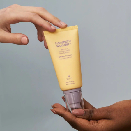 Deux mains tiennent un tube jaune de crème solaire Haruharu Wonder sur fond gris clair. Le tube porte un texte précisant sa protection SPF 50+. Dakar