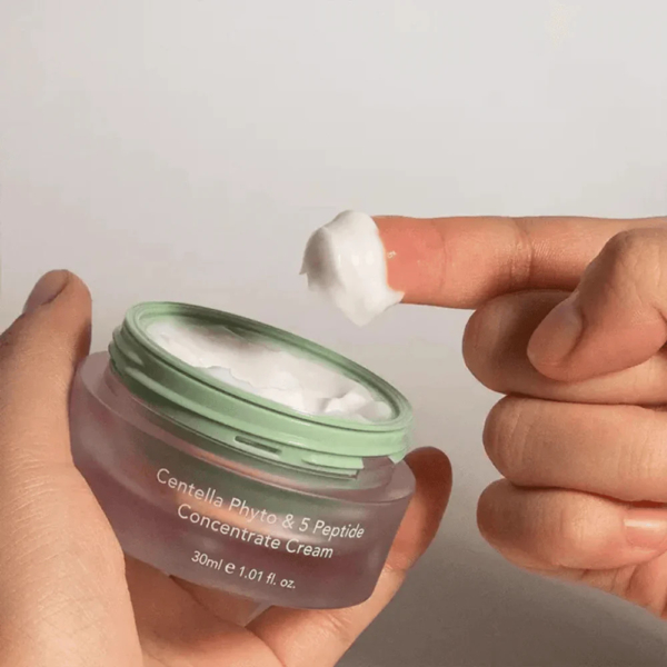 Une personne tient un pot ouvert de crème Centella Phyto & 5 Peptides Concentrate, avec un couvercle vert. Une autre main trempe le bout d'un doigt dans la crème blanche, révélant sa texture épaisse. L'étiquette indique une contenance de 30 ml. Dakar