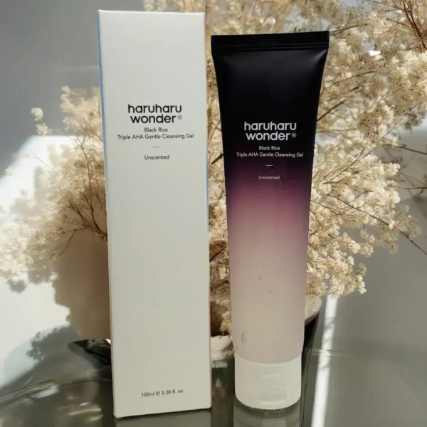 Un tube et une boîte de gel nettoyant doux Haruharu Wonder Black Rice Triple AHA sont présentés sur une surface réfléchissante. Le tube passe du noir au blanc. Des fleurs séchées beiges en arrière-plan apportent une touche d'élégance. Dakar
