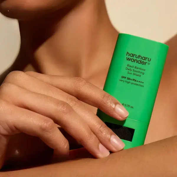 Gros plan d'une personne tenant contre son épaule un tube vert de crème solaire apaisante quotidienne Haruharu Wonder Black Bamboo SPF 50+/PA++++. L'épaule de la personne est nue et sa main saisit délicatement le produit. Dakar
